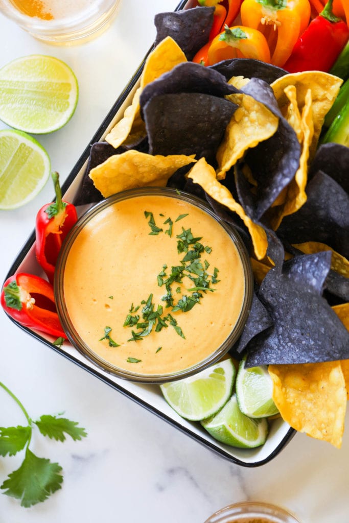A black rimmed tray filled with vegan nacho cheese in a bowl surrounded with blue and yellow tortillas and fresh cut veggies