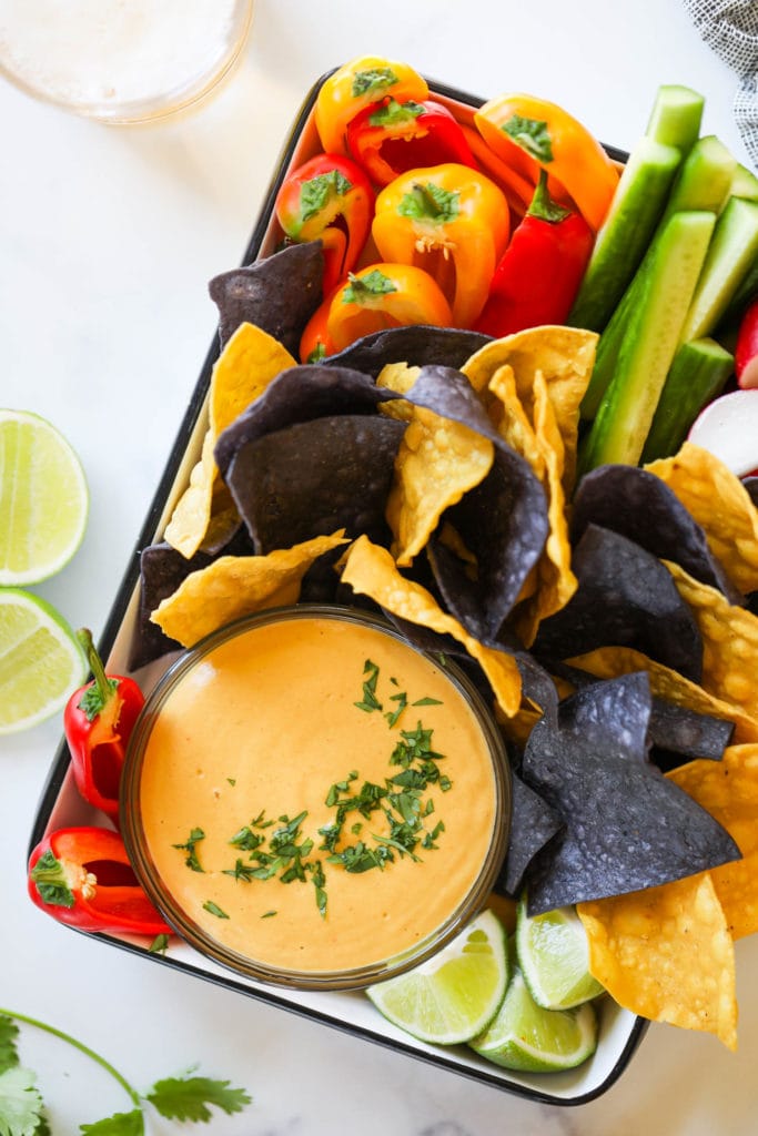 A tray filled with vegan nacho cheese in a bowl surrounded with blue and yellow tortilla chips and fresh cut veggies.