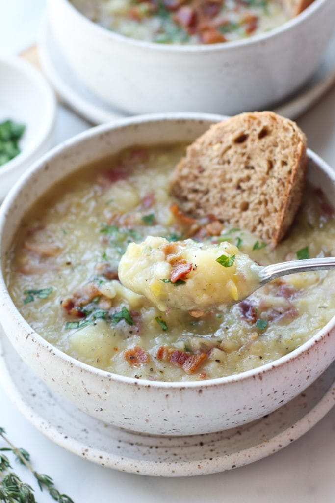 A spoonful of creamy potato leek soup topped with bacon crumbles with crusty bread on the side.