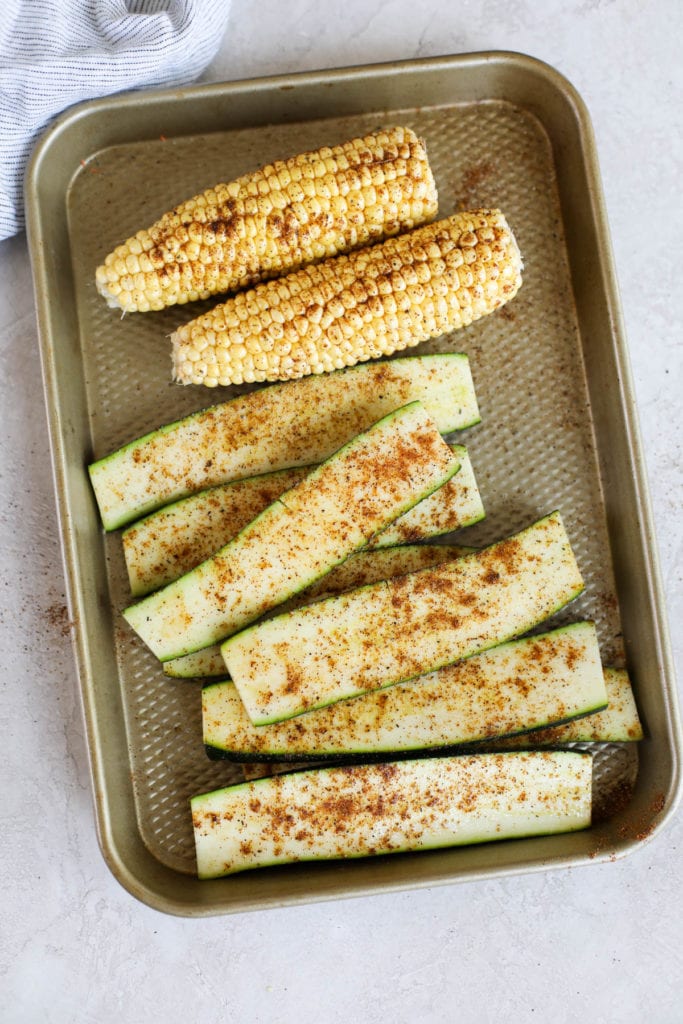 A sheet pan with sliced zucchini and corn on the cob that have been seasoned with chili powder and cumin before grilling.
