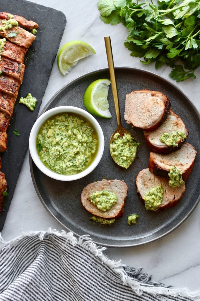 Photo of Grilled Pork Tenderloin sliced, topped with avocado green sauce and served on a plate with a white bowl holding avocado green sauce.