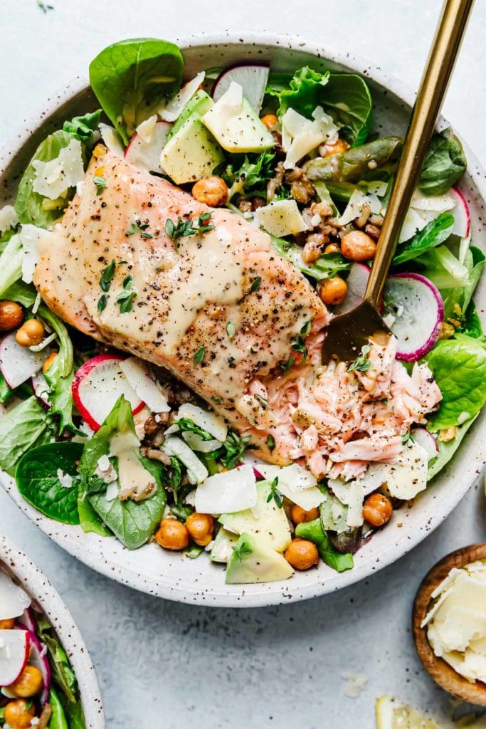 Caesar Salmon Salad Grain Bowl in a speckled stone bowl with greens, avocado, crispy chickpeas and flaky salmon.