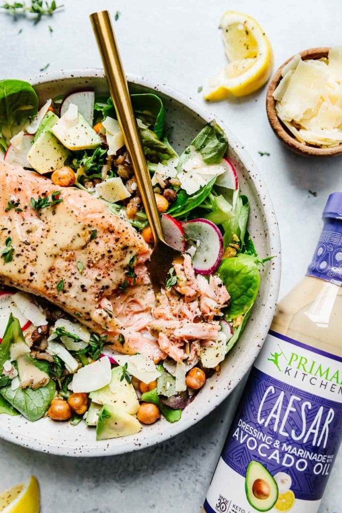 Caesar Salmon Salad Grain Bowl in a speckled stone bowl with greens, avocado, crispy chickpeas, flaky salmon and a bottle of caesar dressing.