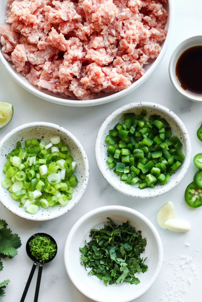Ingredients for pork burgers including green onions, green peppers, jalapenos, and fresh herbs in small bowls.
