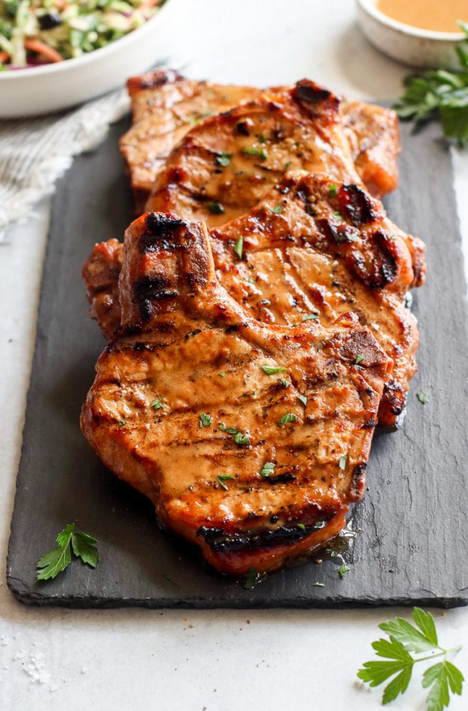 Four marinated grilled pork chops with a homemade honey mustard dressing resting on a black slate board