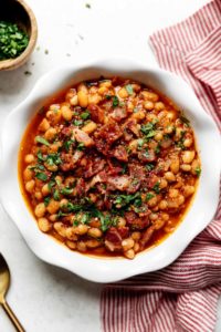 A white scalloped bowl filled with Instant Pot Baked Beans and topped with bacon and herbs.