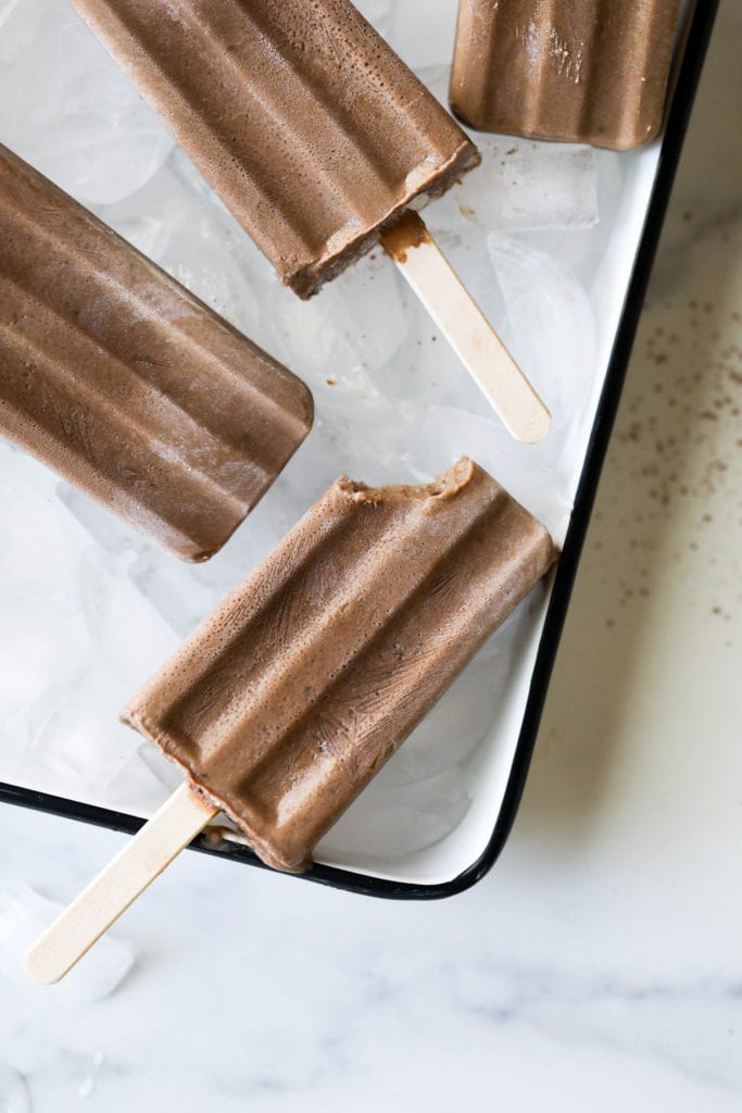 Creamy chocolate dairy-free fudgesicles laying on ice in a try with one fudge pop having a bite taken out of the top edge.