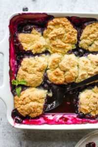 Gluten-free Blueberry Cobbler being scooped out of a white baking dish with a black serving spoon