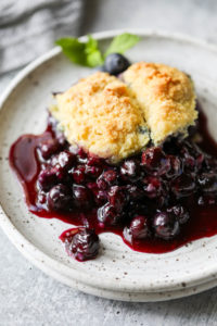Juicy and vibrant dark purple blueberry cobbler topped with a gluten-free biscuit served on a speckled plate