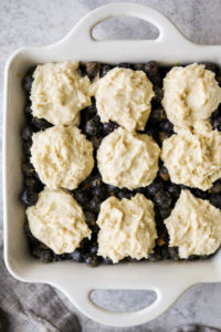 Gluten-free biscuit dollops on top of fresh blueberries in a white baking dish