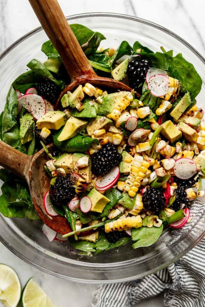 Grilled corn salad being tossed with wooden spoons to mix together blackberries, avocado, radishes, and spinach.