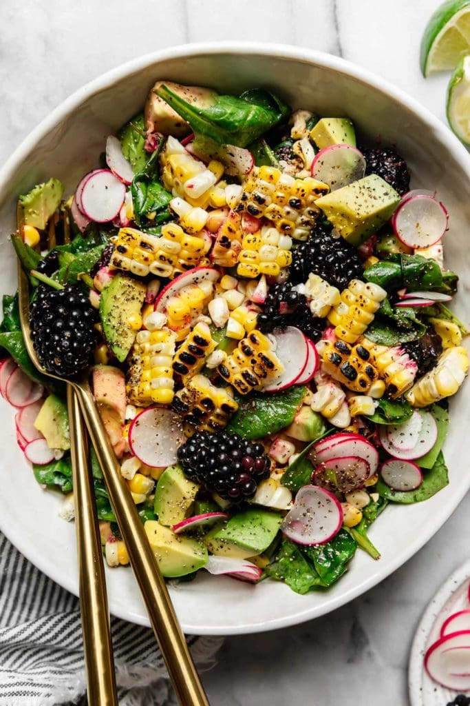 Overhead view of grilled corn salad with avocado, blackberries, and thinly sliced radishes in a white bowl with gold cutlery.