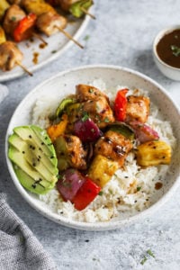 Teriyaki chicken with pineapple and peppers served over rice plated on a speckled plate with sliced avocado on the side