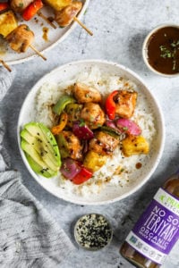 Grilled chicken kebabs with pineapple, peppers, and red onion over rice with a side of sliced avocado plated 